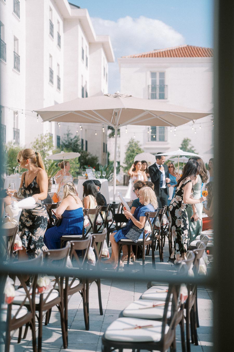 Outdoor wedding reception with guests mingling and sitting under umbrellas, set against a backdrop of white buildings.