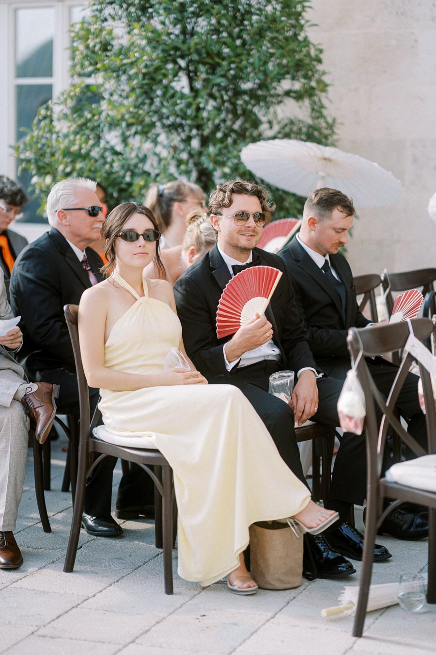 Guests sitting outdoors at a formal event, wearing elegant attire and sunglasses, some holding red fans and white umbrellas for shade.