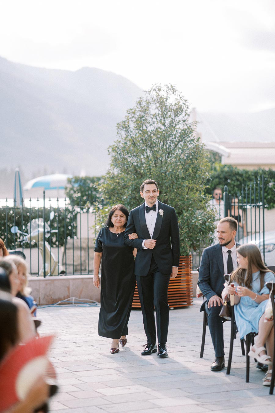 Elegant wedding scene with a well-dressed man in a tuxedo and a woman in a formal black dress walking arm in arm down an outdoor aisle, surrounded by seated guests.
