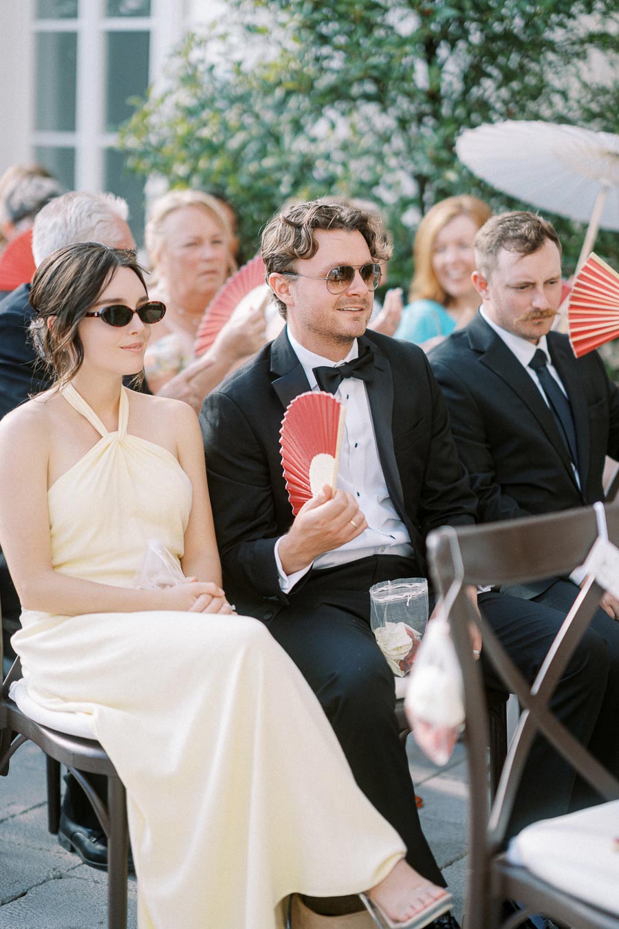 Guests seated at an outdoor wedding ceremony, dressed in formal attire, using red handheld fans to keep cool.