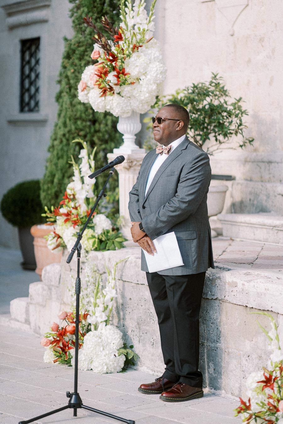 A well-dressed man in a suit and bow tie stands with a microphone in front of an elegant stone building adorned with vibrant floral arrangements, preparing to speak at an outdoor event.