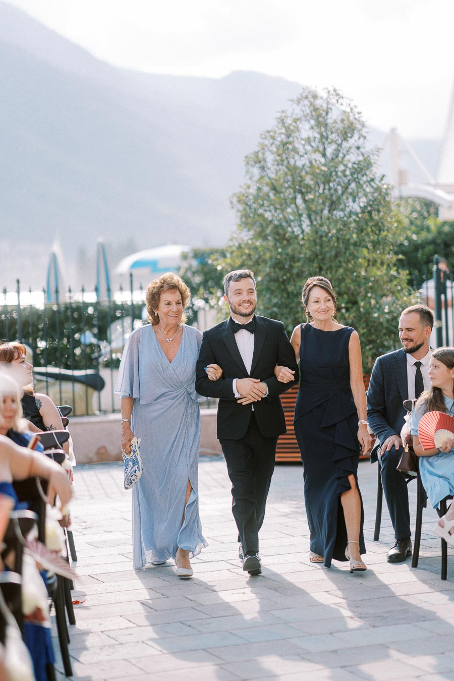 Elegant outdoor wedding ceremony with guests in formal attire, including a man in a tuxedo escorting two women in evening gowns, against a scenic backdrop.
