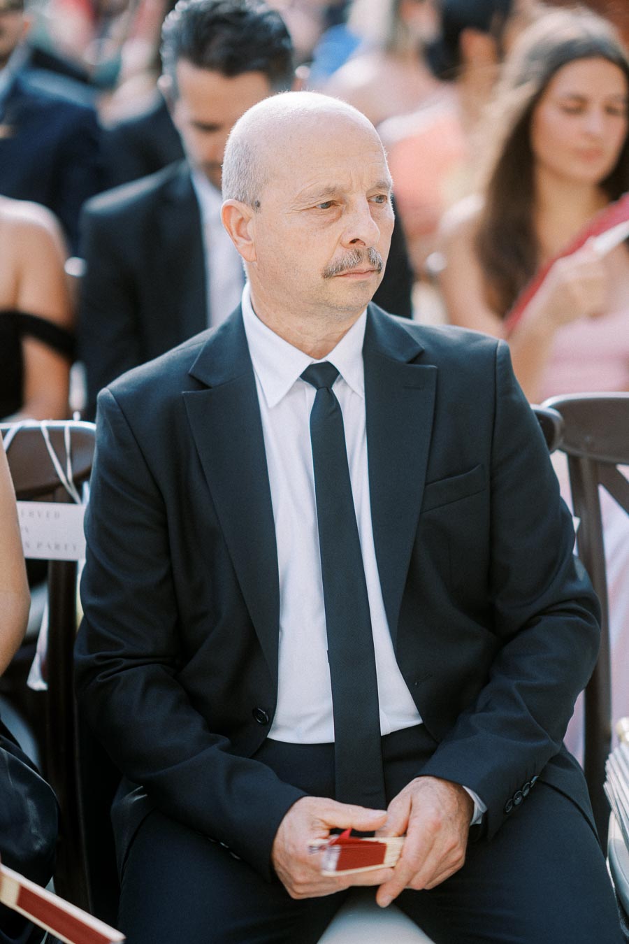 Man in a formal black suit seated outdoors at an event, holding a small item in his hands, surrounded by other attendees with blurred background.