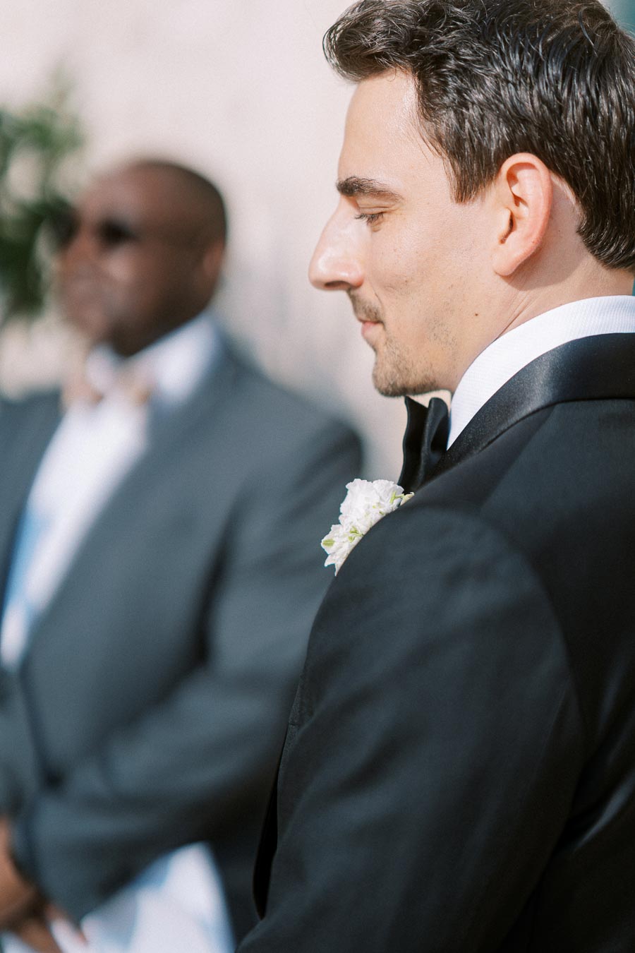 Groom in profile wearing a black tuxedo with a white floral boutonniere, standing alongside an attendee in a blurred background, at an outdoor wedding ceremony.