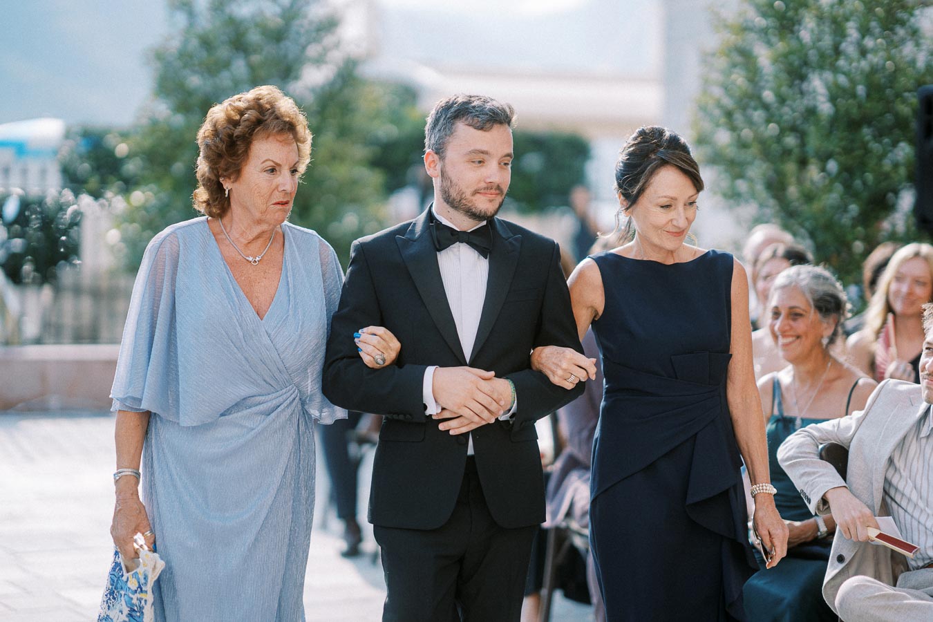 Man in tuxedo escorts two women in elegant dresses during outdoor wedding ceremony, with smiling guests in background