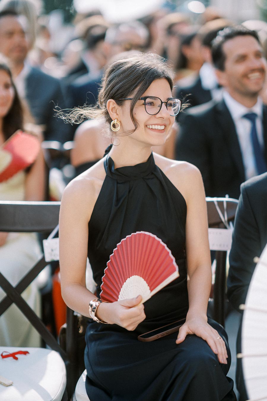 A woman in a black dress and glasses smiling while holding a red hand fan at an outdoor event.