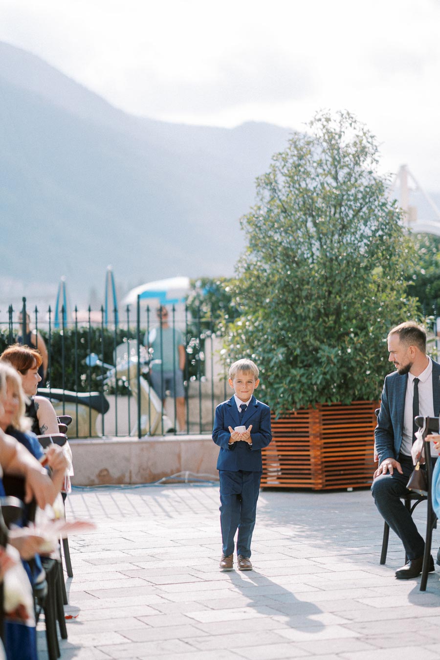 A young boy dressed in a blue suit walks down an outdoor aisle during a sunny wedding ceremony, holding a small white pillow as guests watch from either side.