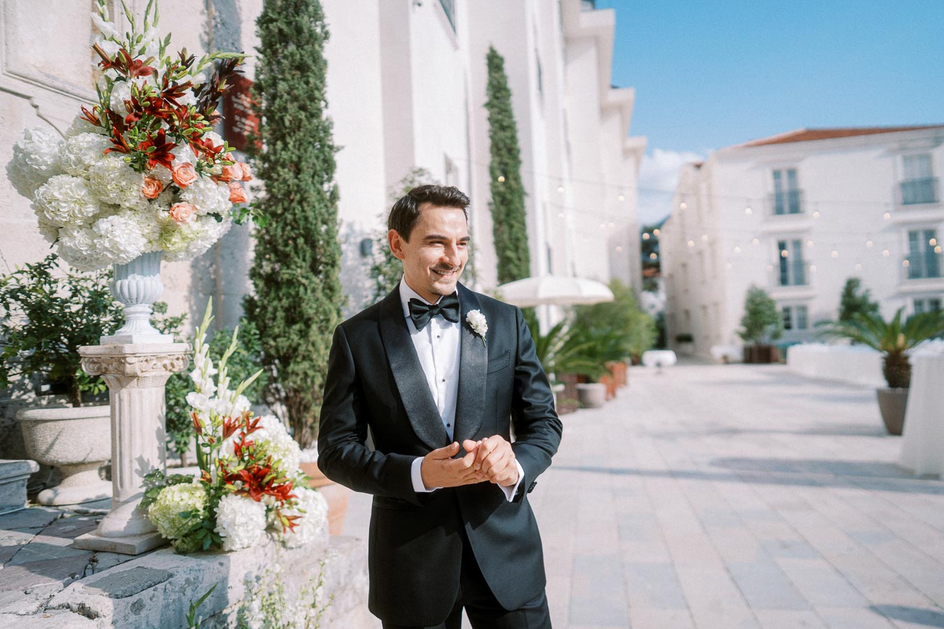 Smiling groom in a black tuxedo standing outdoors with elegant floral arrangements, white stone buildings, and clear blue sky in a picturesque wedding venue.