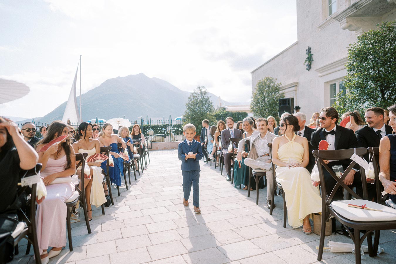 Young boy walking down an outdoor wedding aisle, surrounded by seated guests dressed in formal attire, with picturesque mountain scenery in the background.