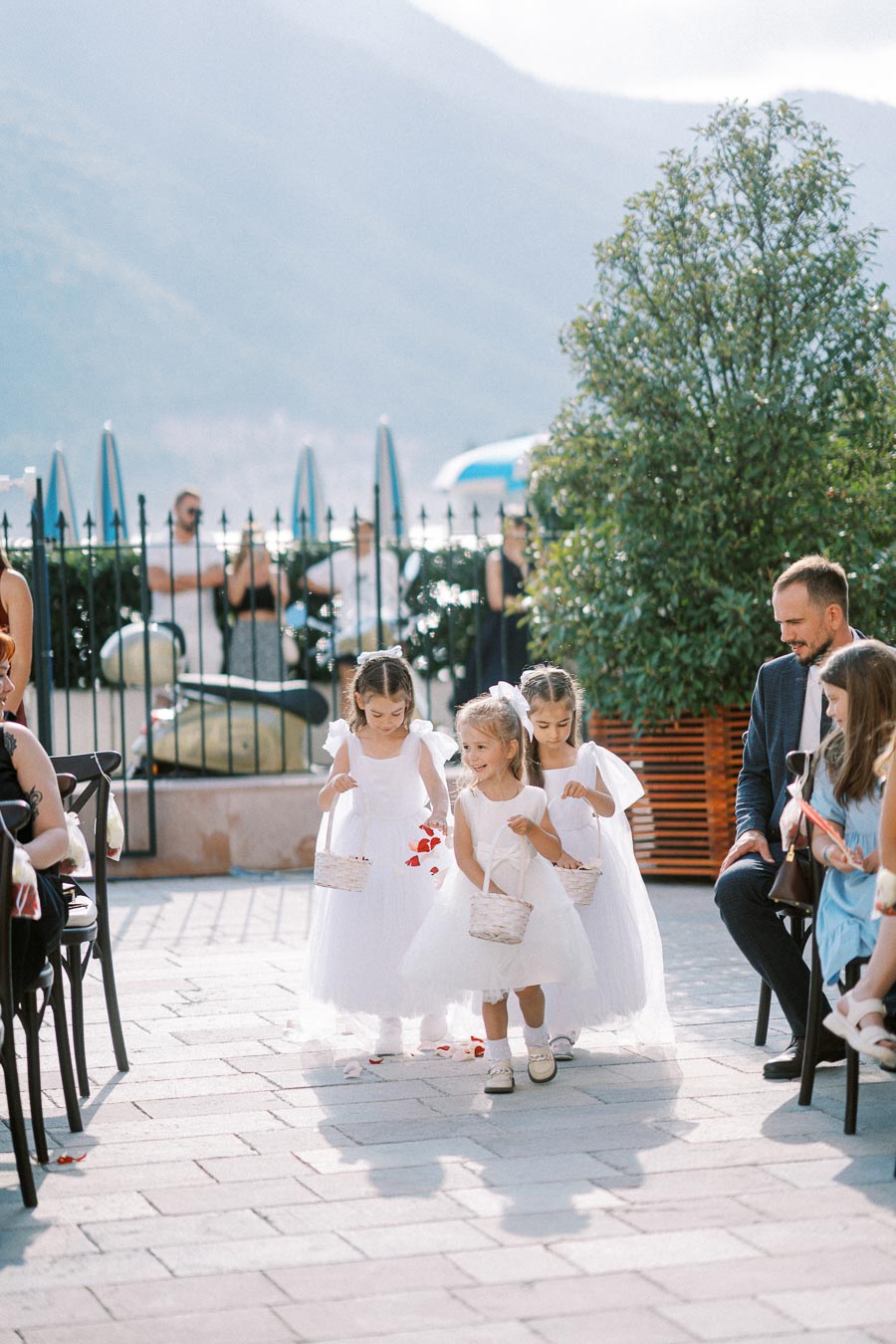 Four young flower girls in white dresses, carrying baskets of petals, walking down a stone pathway during an outdoor wedding ceremony. Guests seated along the aisle, with mountains and a clear sky in the background, create a picturesque setting.