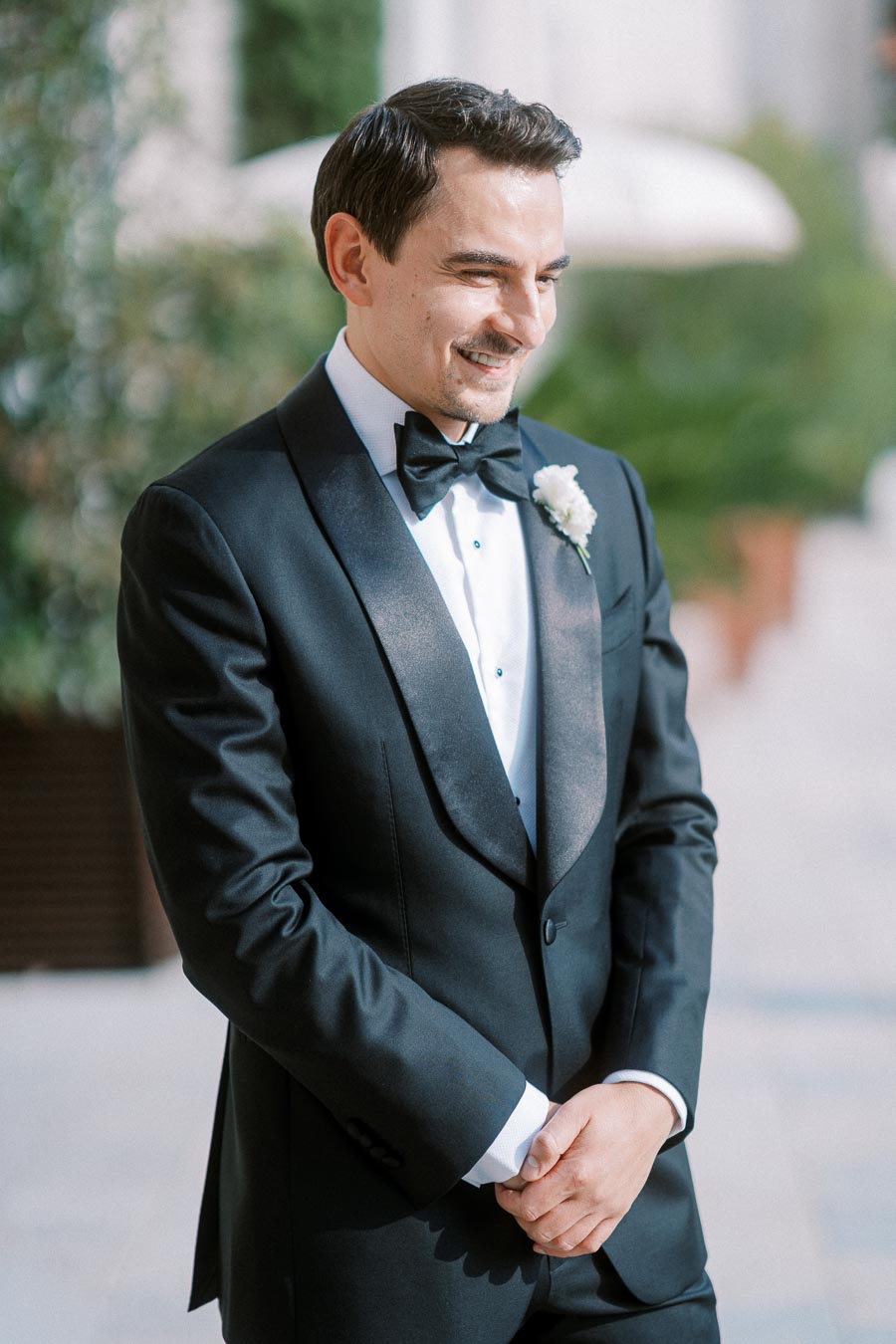 Elegant groom in a black tuxedo and bow tie smiling warmly outdoors.