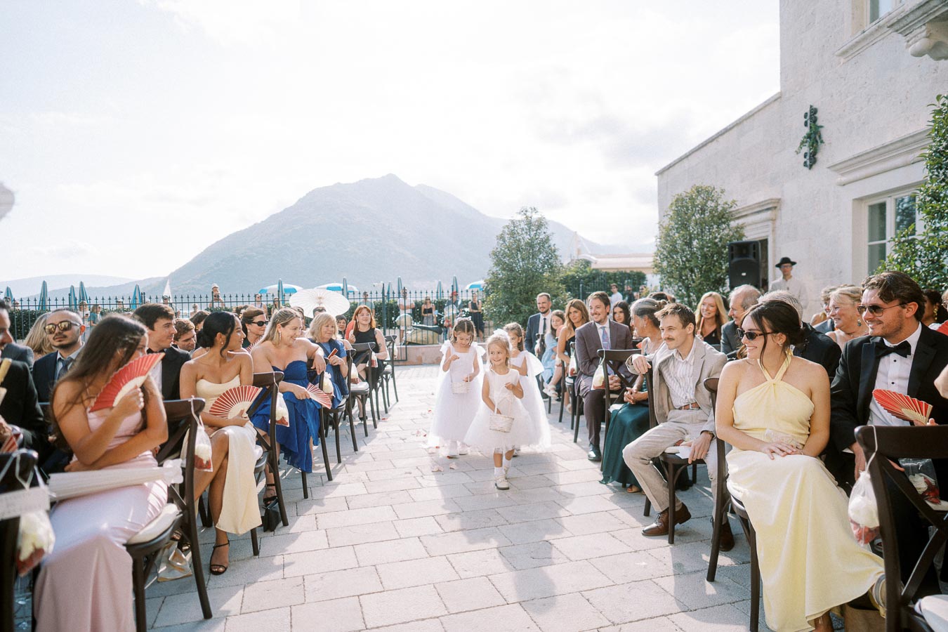 Outdoor wedding ceremony with guests seated along an aisle, featuring flower girls walking in white dresses; scenic mountain backdrop under a bright sky.