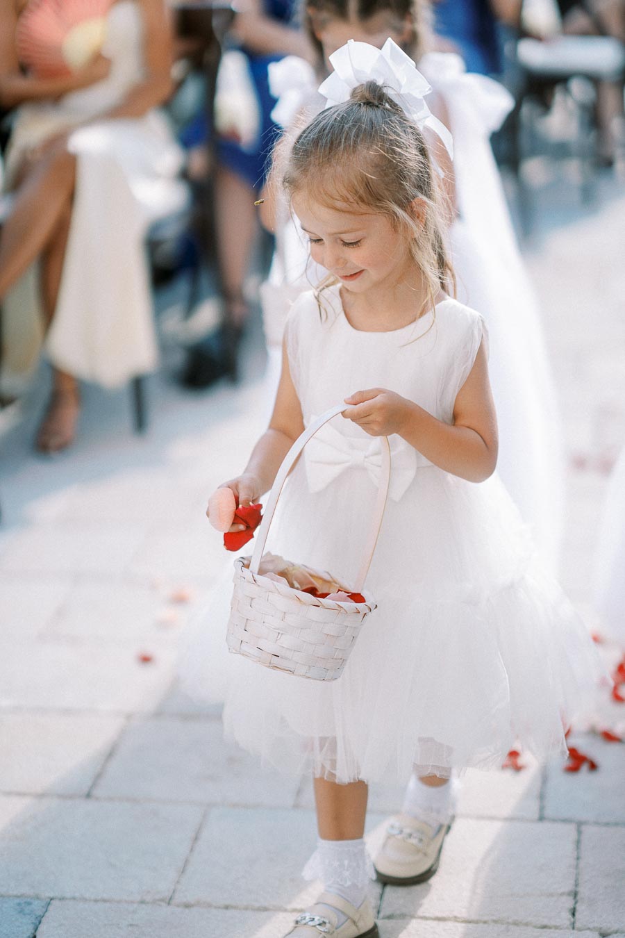 A young flower girl in a white dress and bow carrying a basket of red petals during a wedding ceremony.