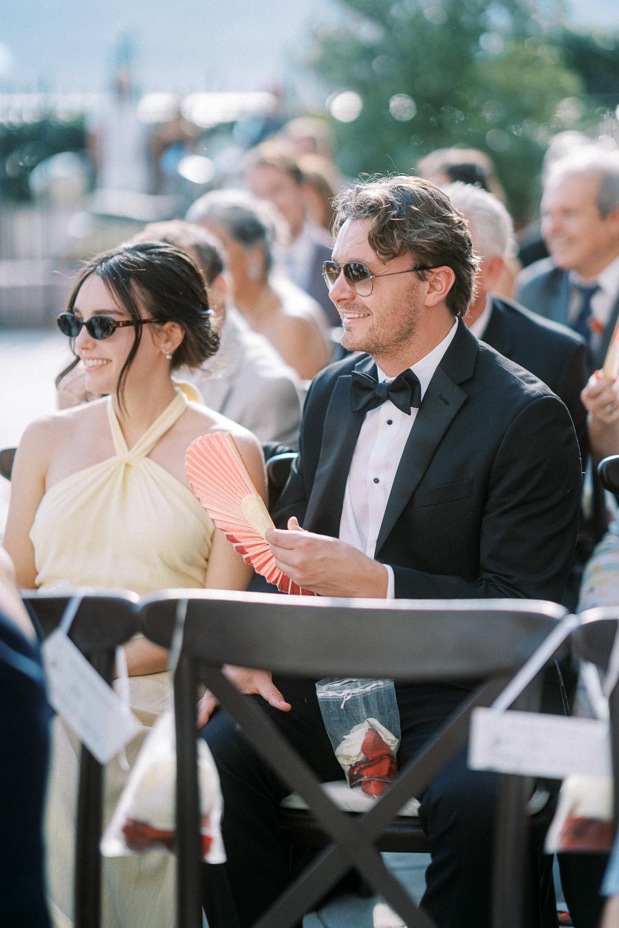 Guests at a wedding ceremony sitting outdoors, wearing formal attire and sunglasses, holding a decorative fan.