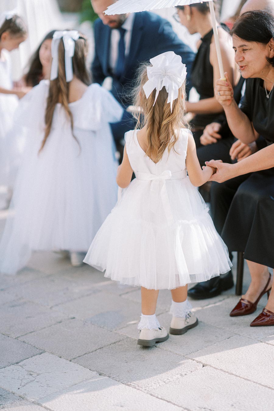A young girl wearing a white dress with a large bow stands holding hands with an adult woman under a parasol at a wedding ceremony. Other children in white dresses are visible in the background, adding to the festive atmosphere.