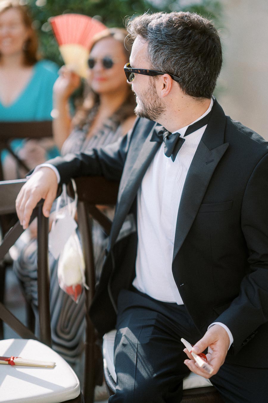 A man in a black tuxedo and sunglasses sitting on a chair at an outdoor event, with two women in the background, one holding a red and yellow fan, creating a lively and elegant atmosphere.