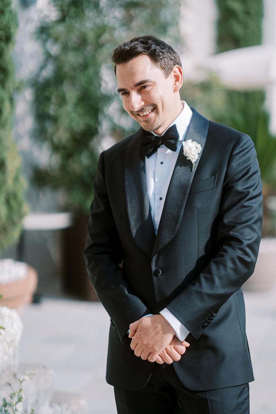 Smiling groom in a black tuxedo with a white boutonniere, standing outdoors on a sunny day.