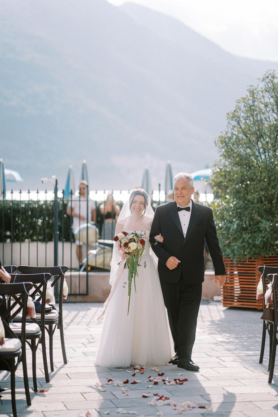 Bride walking down the aisle with her father at an outdoor wedding ceremony, holding a bouquet of red and white flowers, with a scenic mountain backdrop.