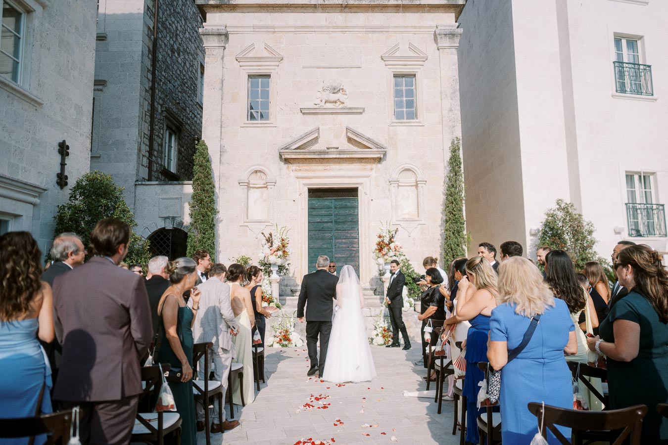 A bride and groom standing at the altar during an outdoor wedding ceremony, surrounded by guests in formal attire, with a historic stone building in the background.