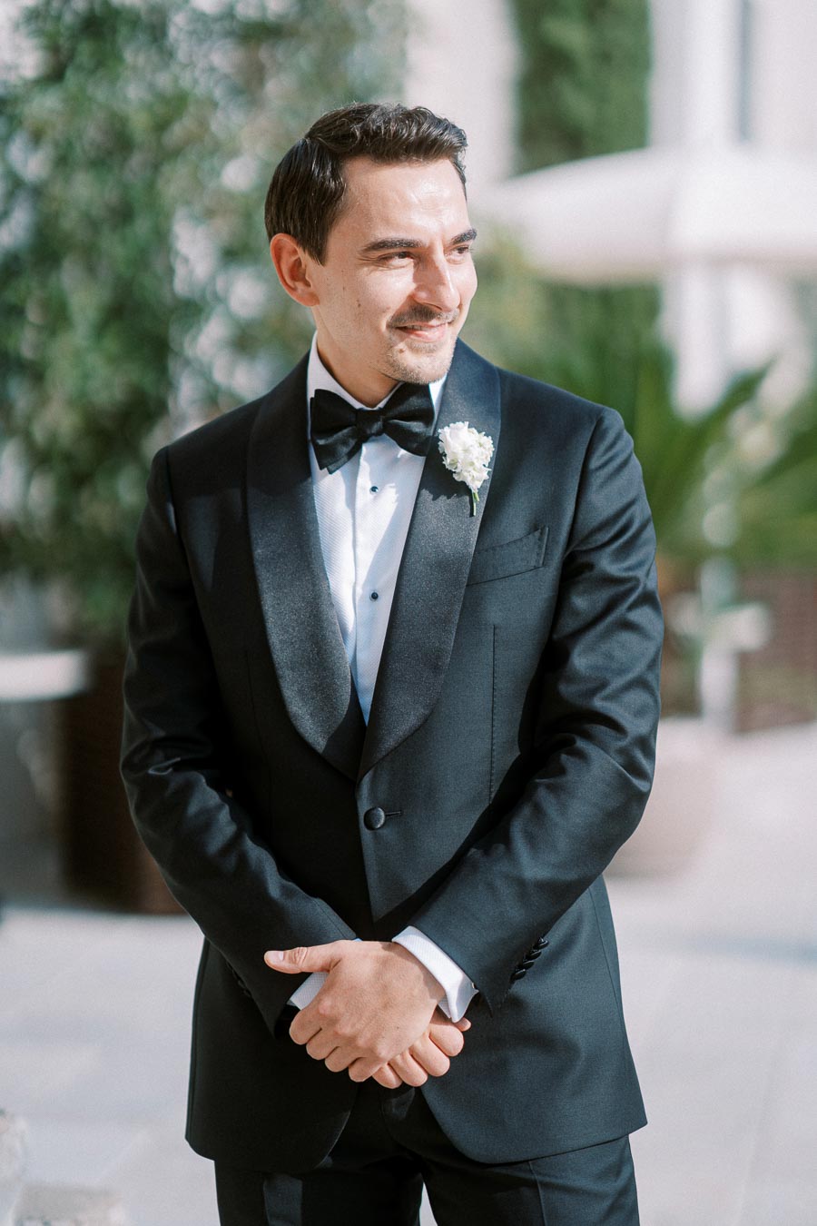 Elegant man in a tuxedo with a bow tie and boutonnière stands outdoors, smiling and posing for a formal occasion.