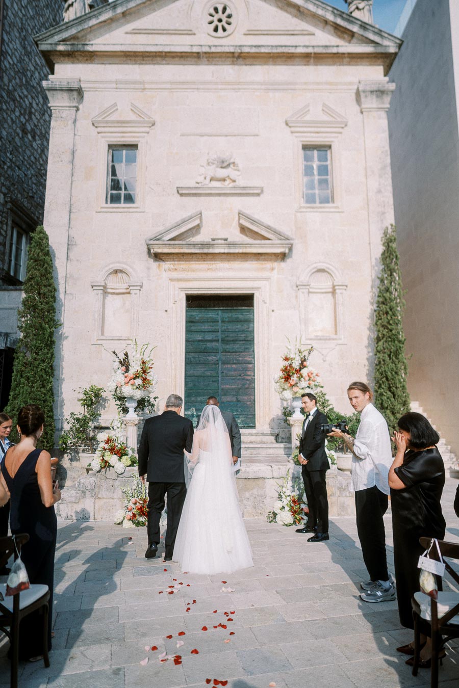 A wedding ceremony taking place in front of a historic stone church, with a bride in a white gown and veil walking down an aisle adorned with rose petals, accompanied by guests and a photographer capturing the moment.