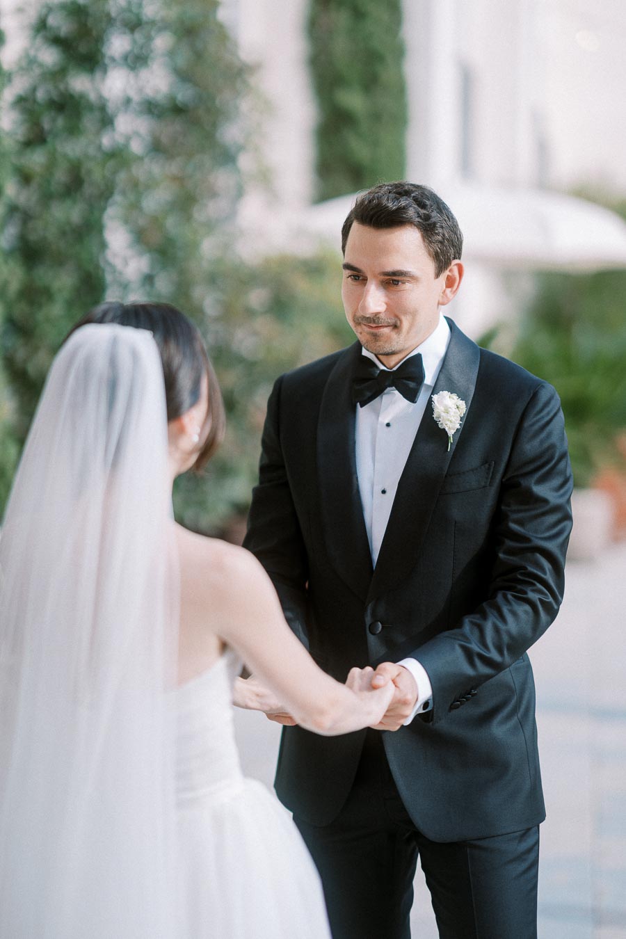 A groom in a black tuxedo holds hands with his bride in a white wedding dress and veil during an outdoor wedding ceremony.