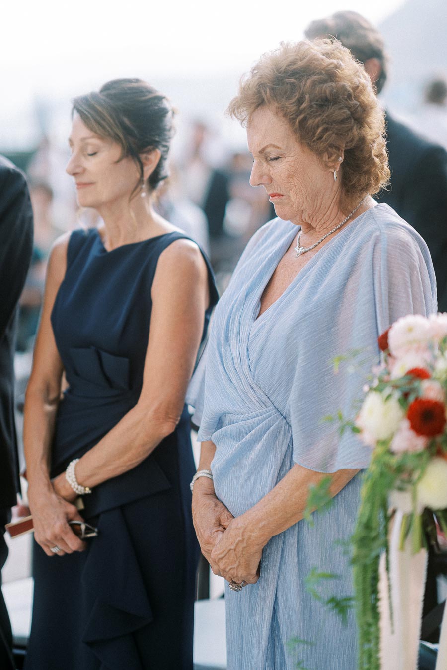 Two elegantly dressed women standing during a formal event, wearing blue outfits with floral arrangements in the foreground.