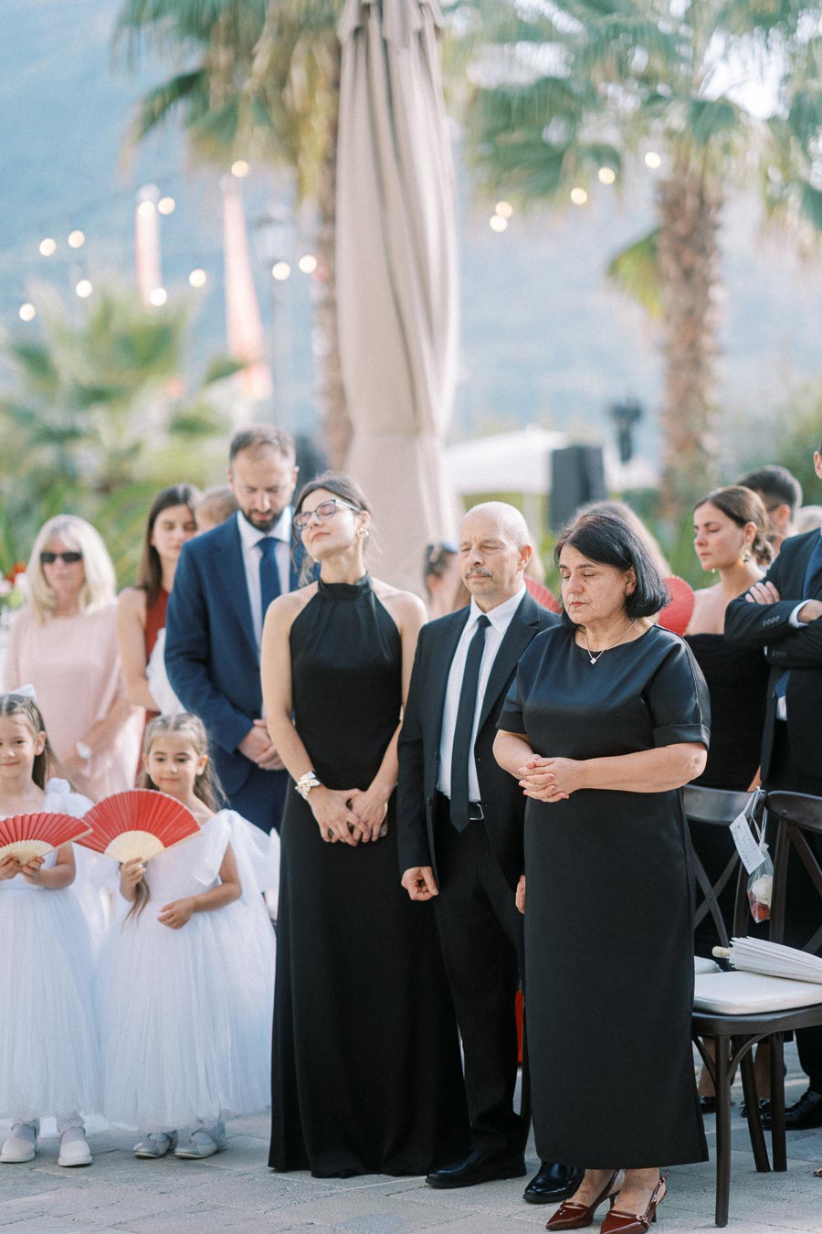 A group of people, including adults in formal attire and young girls in white dresses holding red fans, standing at an outdoor event with palm trees and bokeh lights in the background.