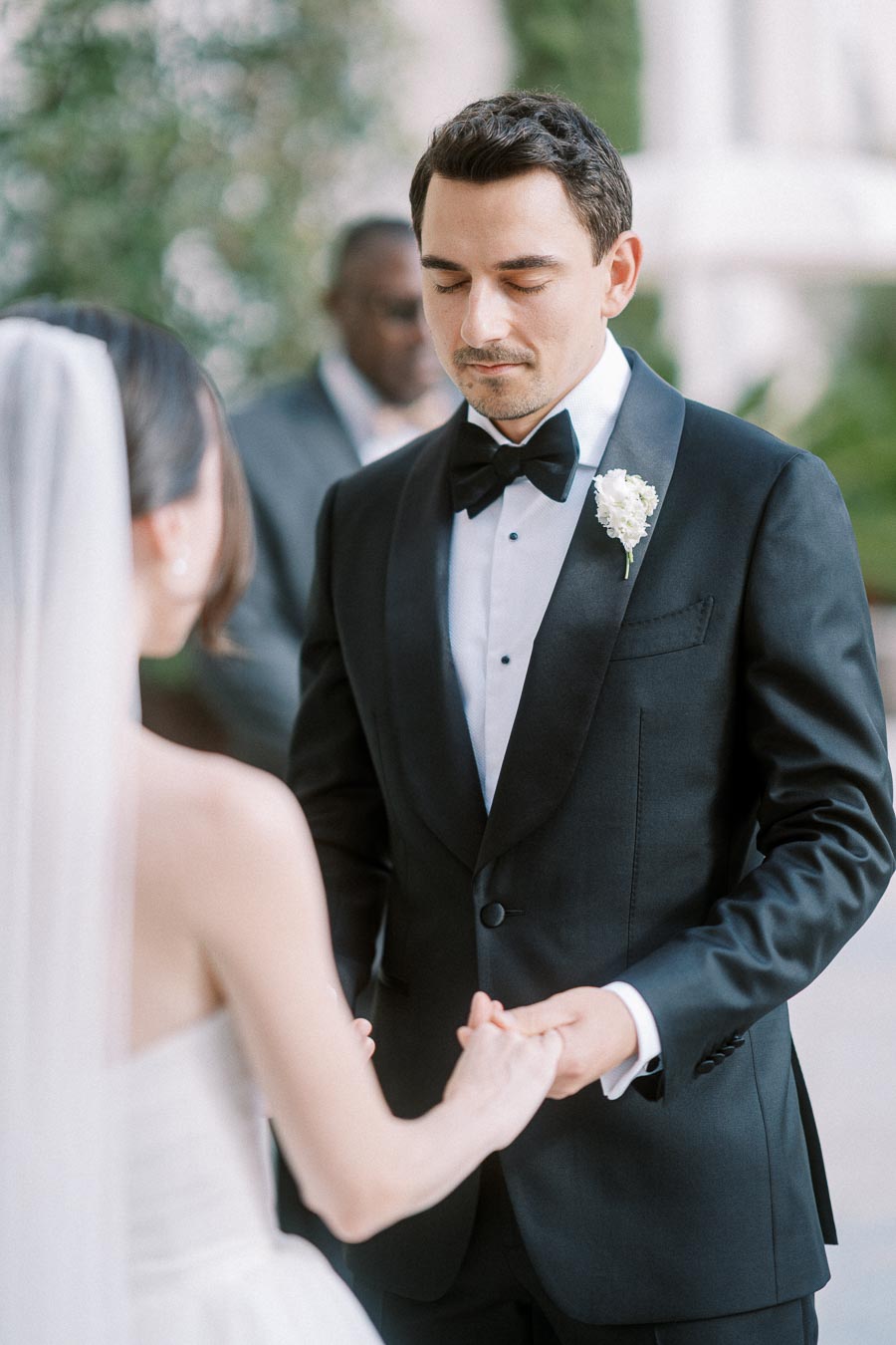 Bride and groom holding hands during wedding ceremony, groom in black tuxedo with white boutonnière.