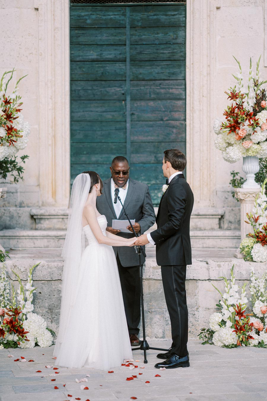 Outdoor wedding ceremony with a bride in a white gown and veil, holding hands with the groom in a black suit. The officiant stands between them in front of a rustic blue door, surrounded by vibrant floral arrangements.