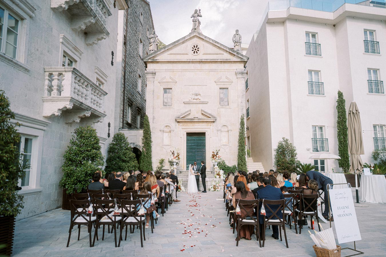 Elegant outdoor wedding ceremony taking place at a historic stone building with guests seated on either side of the aisle adorned with rose petals, creating a romantic and scenic atmosphere for the bride and groom exchanging vows.
