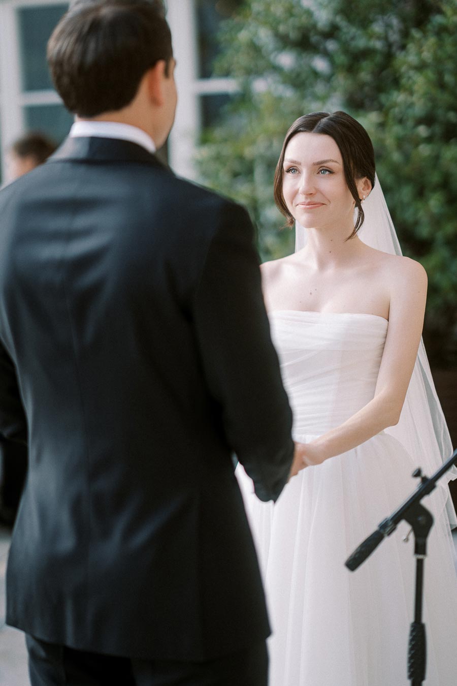 Bride and groom holding hands during an outdoor wedding ceremony, with the bride in a white dress and veil looking at her partner in a black suit.