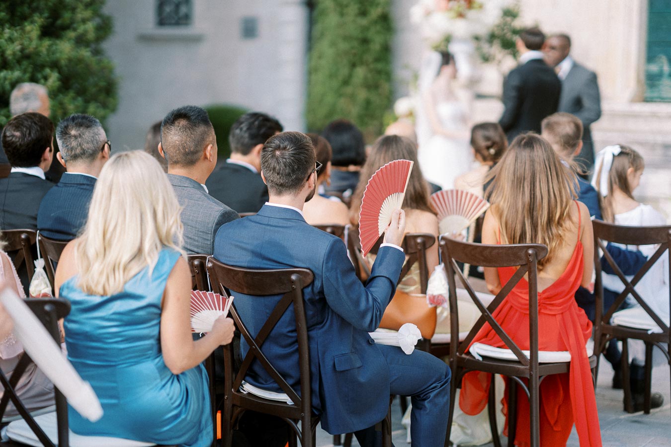 Guests seated at an outdoor wedding ceremony, dressed in formal attire, using paper fans to cool down, with the bride and groom exchanging vows in the background.