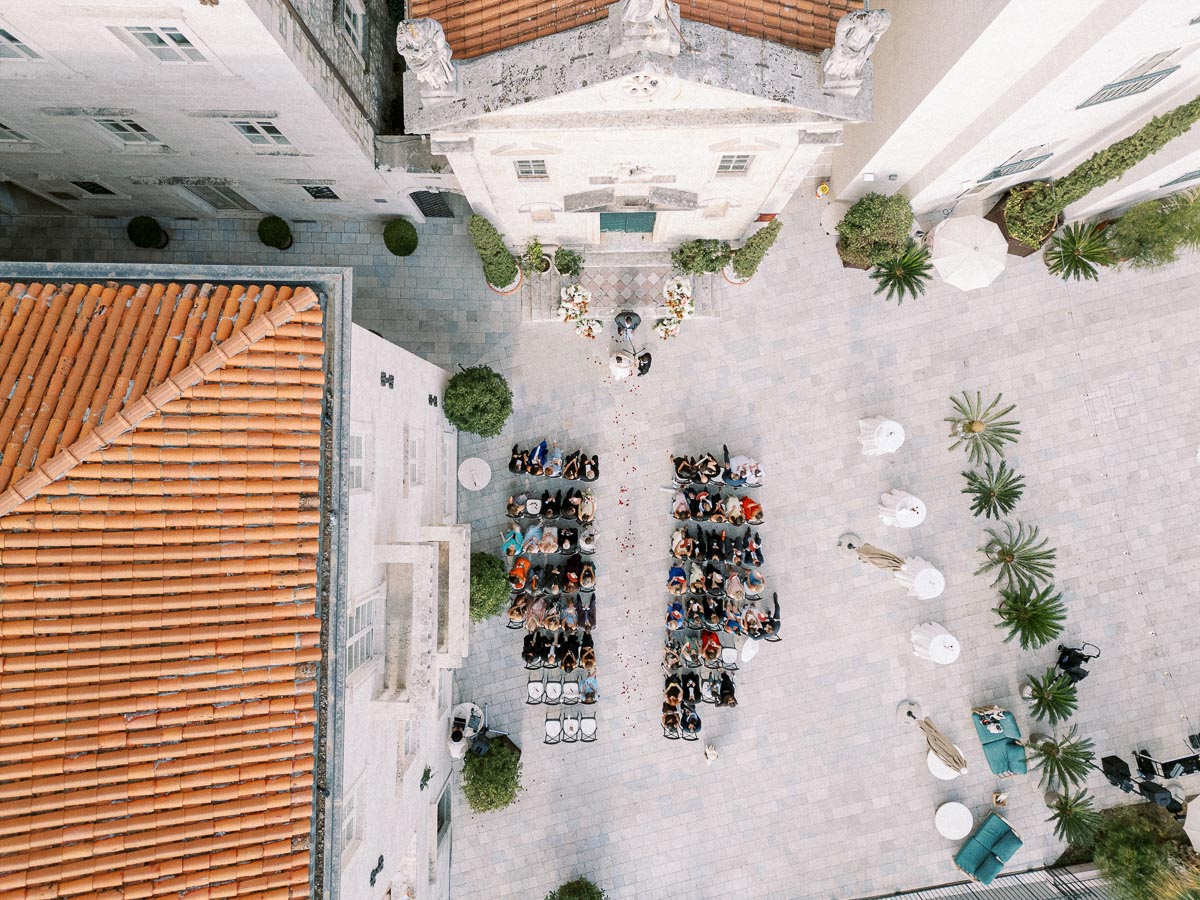 Aerial view of an outdoor wedding ceremony in a historic stone courtyard with guests seated in rows facing an altar adorned with flowers, surrounded by Mediterranean architecture and lush greenery.