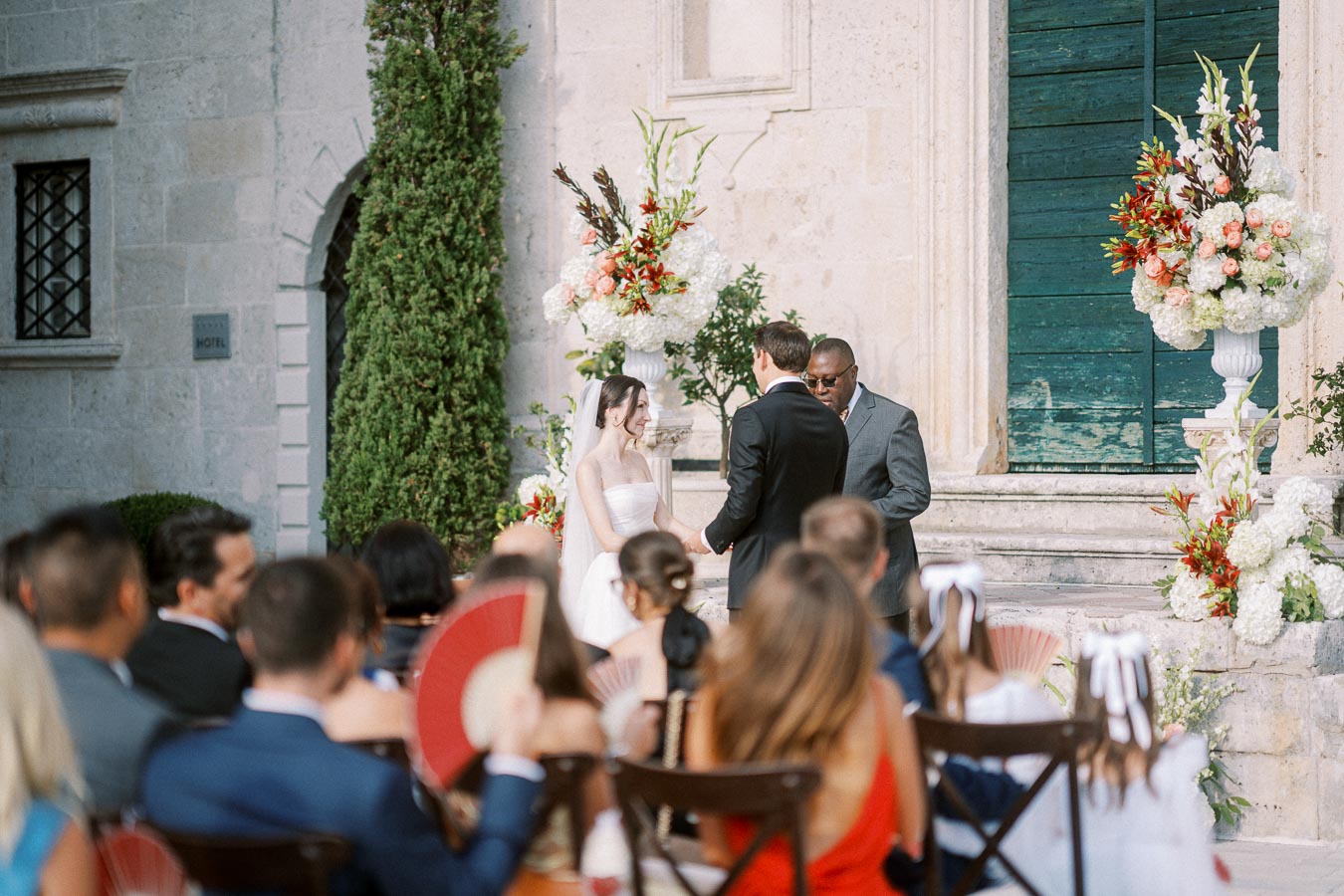 A wedding ceremony taking place outdoors, featuring a bride in a white dress and a groom in a black suit exchanging vows in front of an officiant. The setting includes elegant floral arrangements and seated guests watching the event.