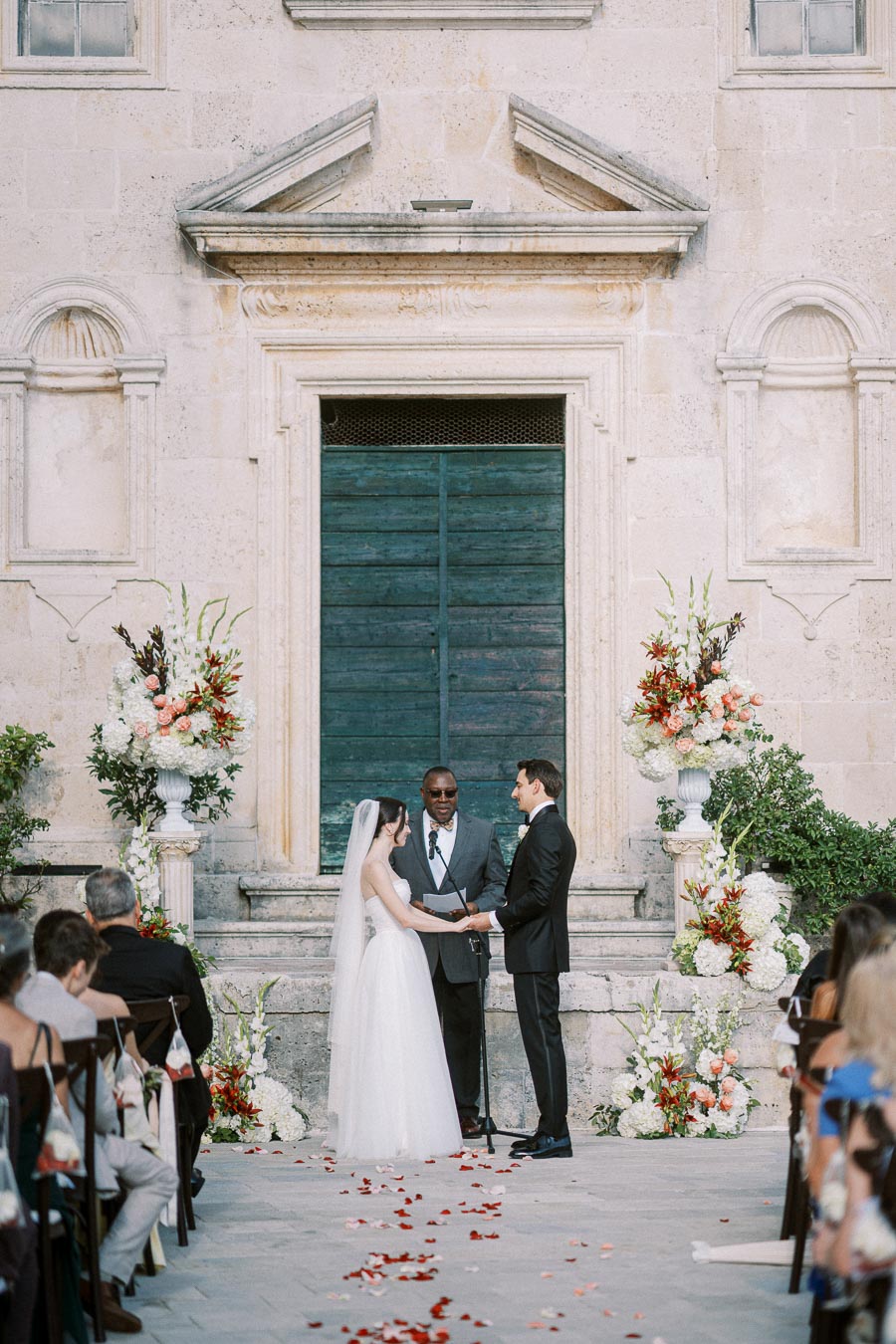 A bride and groom exchange vows during an outdoor wedding ceremony in front of an elegant stone building, with a floral arrangement on either side and guests seated in rows.