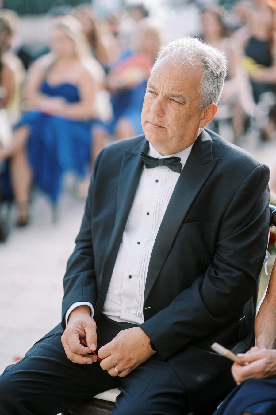 A man in a tuxedo seated at an outdoor event, surrounded by guests in formal attire.