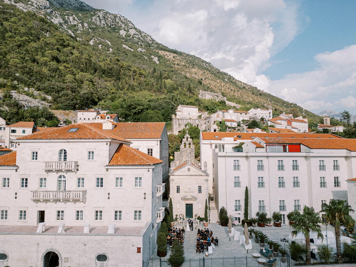 Aerial view of a historic European town with white stone buildings and red-tiled roofs, surrounded by lush green mountains and a partly cloudy sky. A central courtyard hosts an outdoor event with neatly arranged seating, showcasing the charm and architecture typical of Mediterranean regions.