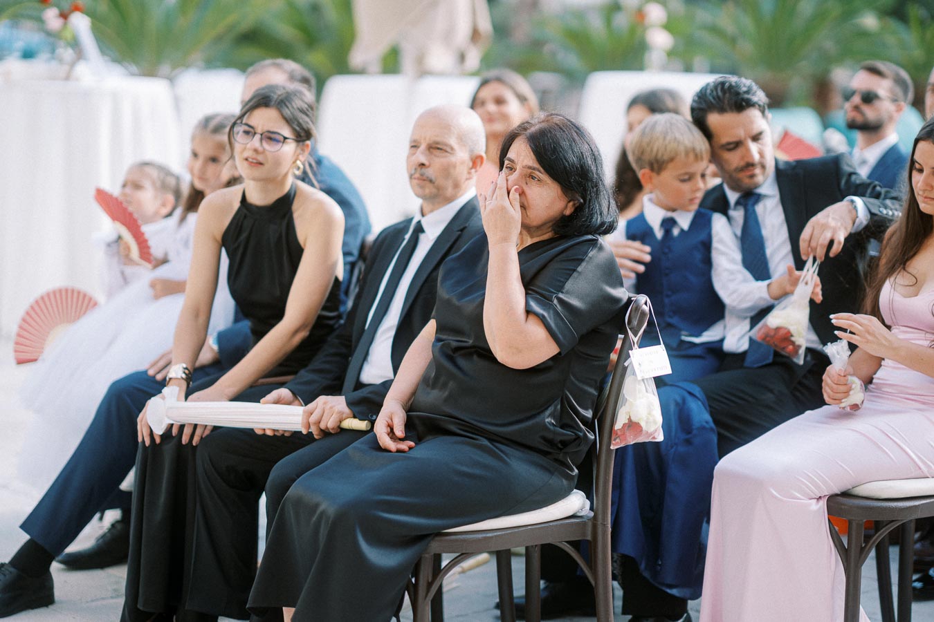 A group of wedding guests seated outdoor; a woman in a black dress appears emotional, surrounded by family and friends, with trees and decorations in the background.