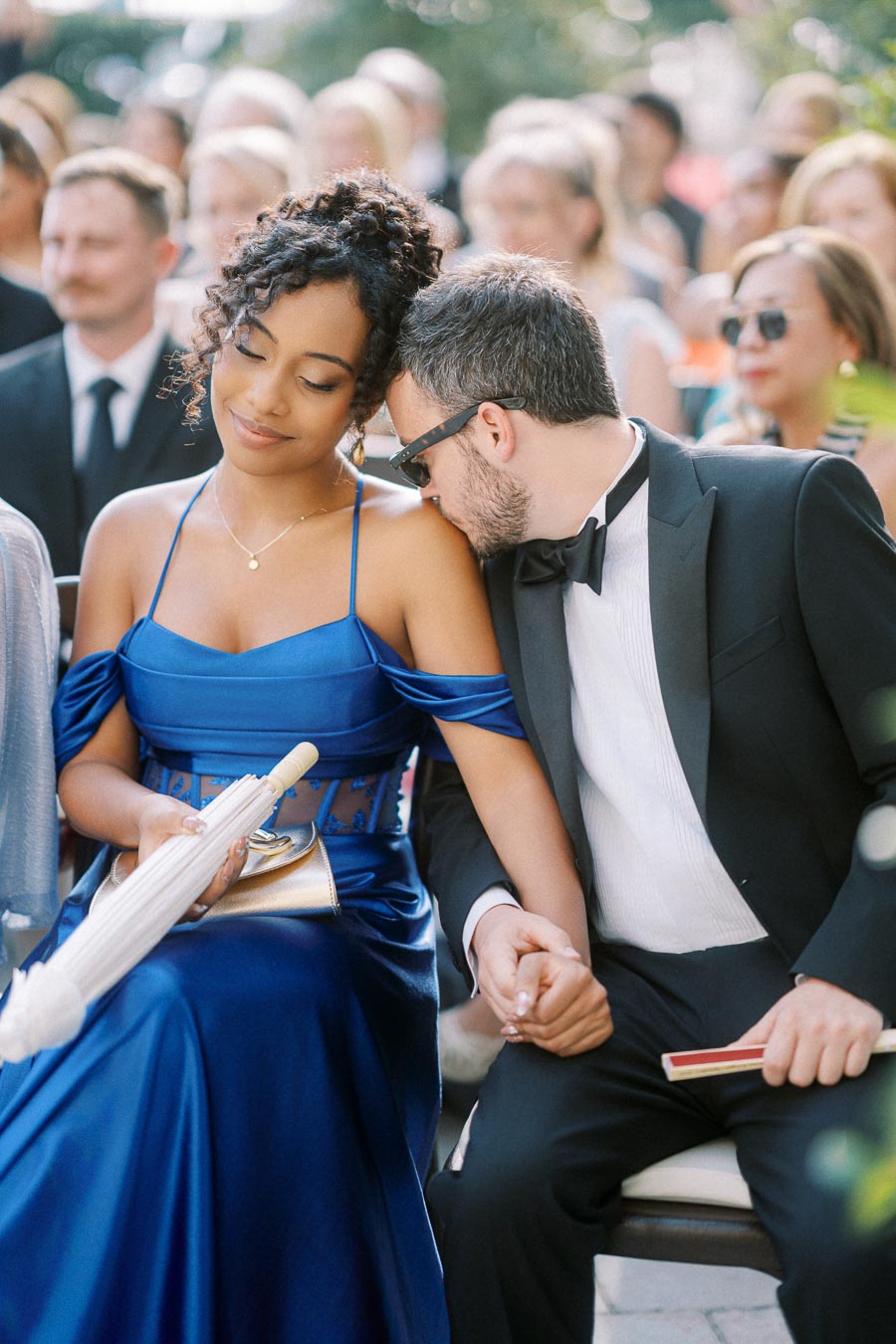 A couple seated together at an outdoor event, the woman in an elegant blue dress holding a parasol, and the man in a black tuxedo and sunglasses, leaning affectionately on her shoulder.