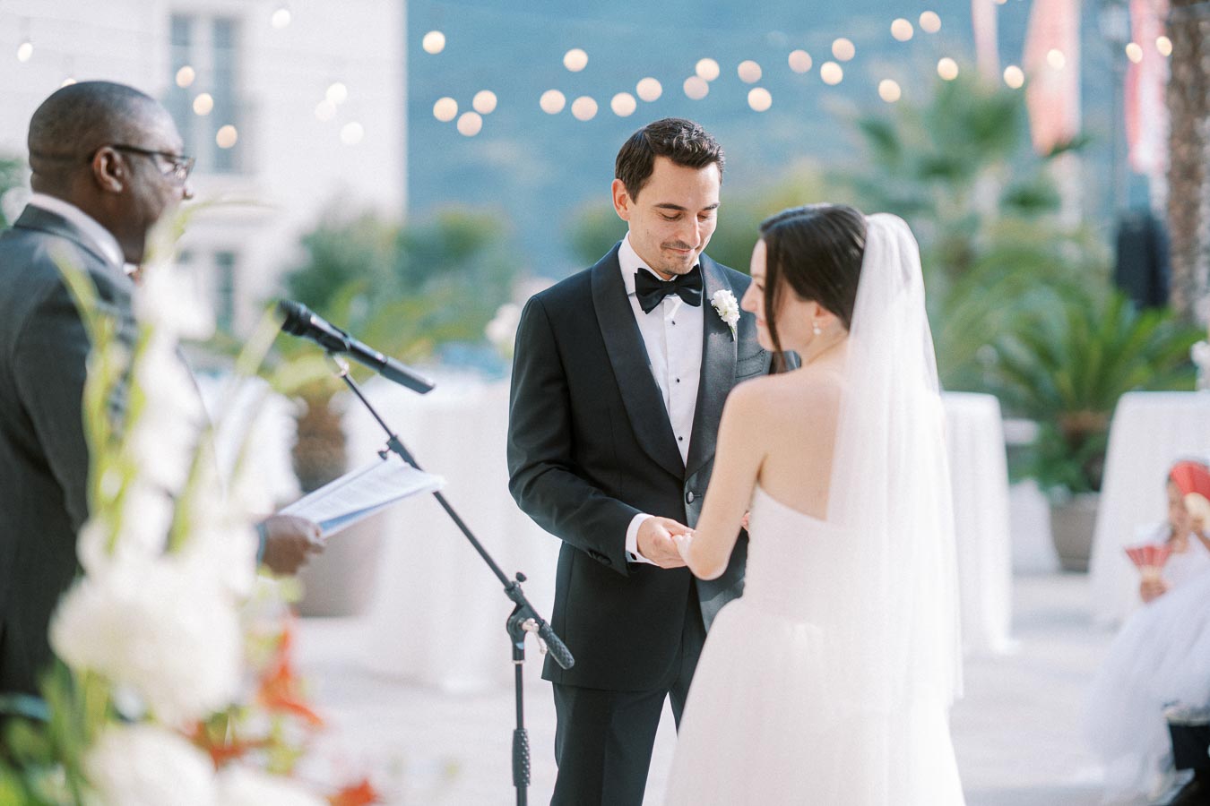 Outdoor wedding ceremony with a groom in a black tuxedo and bride in a white gown exchanging rings, officiated by a man in a suit with a microphone; string lights and scenic background enhance the romantic setting.