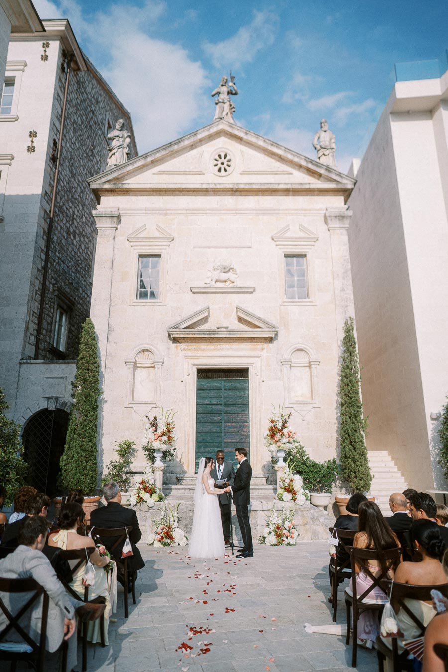 A couple exchanging vows in an outdoor wedding ceremony in front of a historic stone chapel with blue sky above.