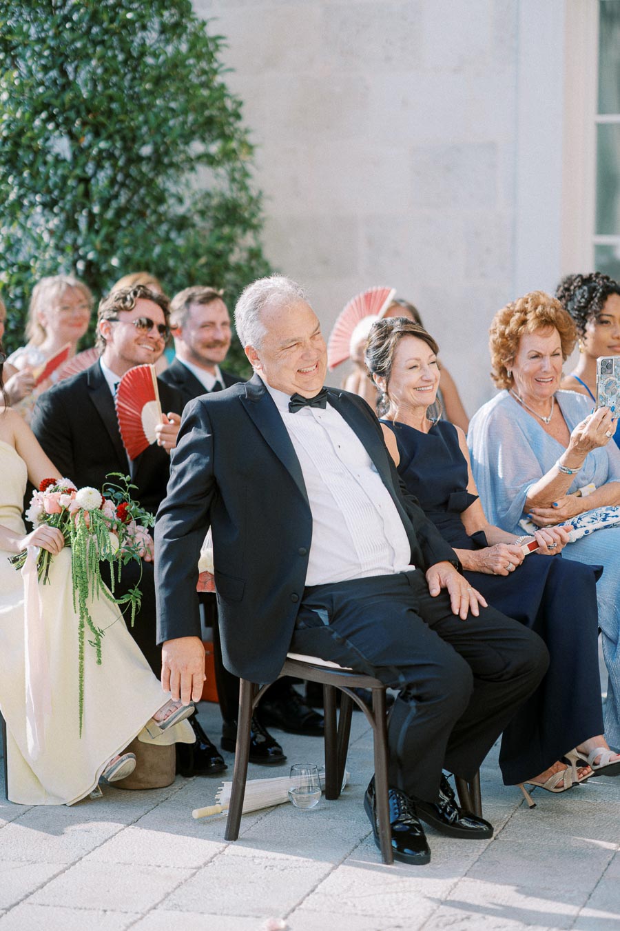 A group of elegantly dressed wedding guests sits outdoors on a sunny day, holding red fans and smiling, with greenery in the background.