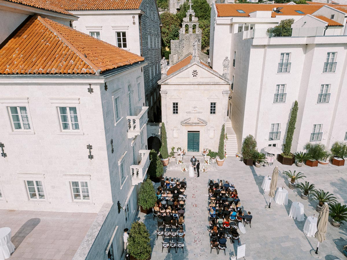 Aerial view of an outdoor wedding ceremony in a historic European courtyard, featuring rows of seated guests, a beautifully decorated altar, and surrounding classic architecture with terracotta roofs.