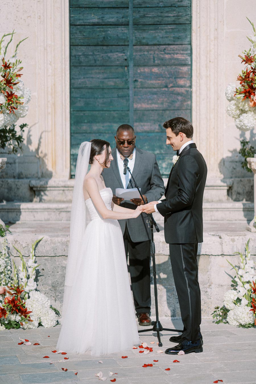 A bride and groom exchange vows during an outdoor wedding ceremony, with an officiant standing between them. The bride is wearing a white gown and veil, while the groom is in a black tuxedo. The scene is adorned with floral arrangements and petals on the ground, set against a decorative stone backdrop.