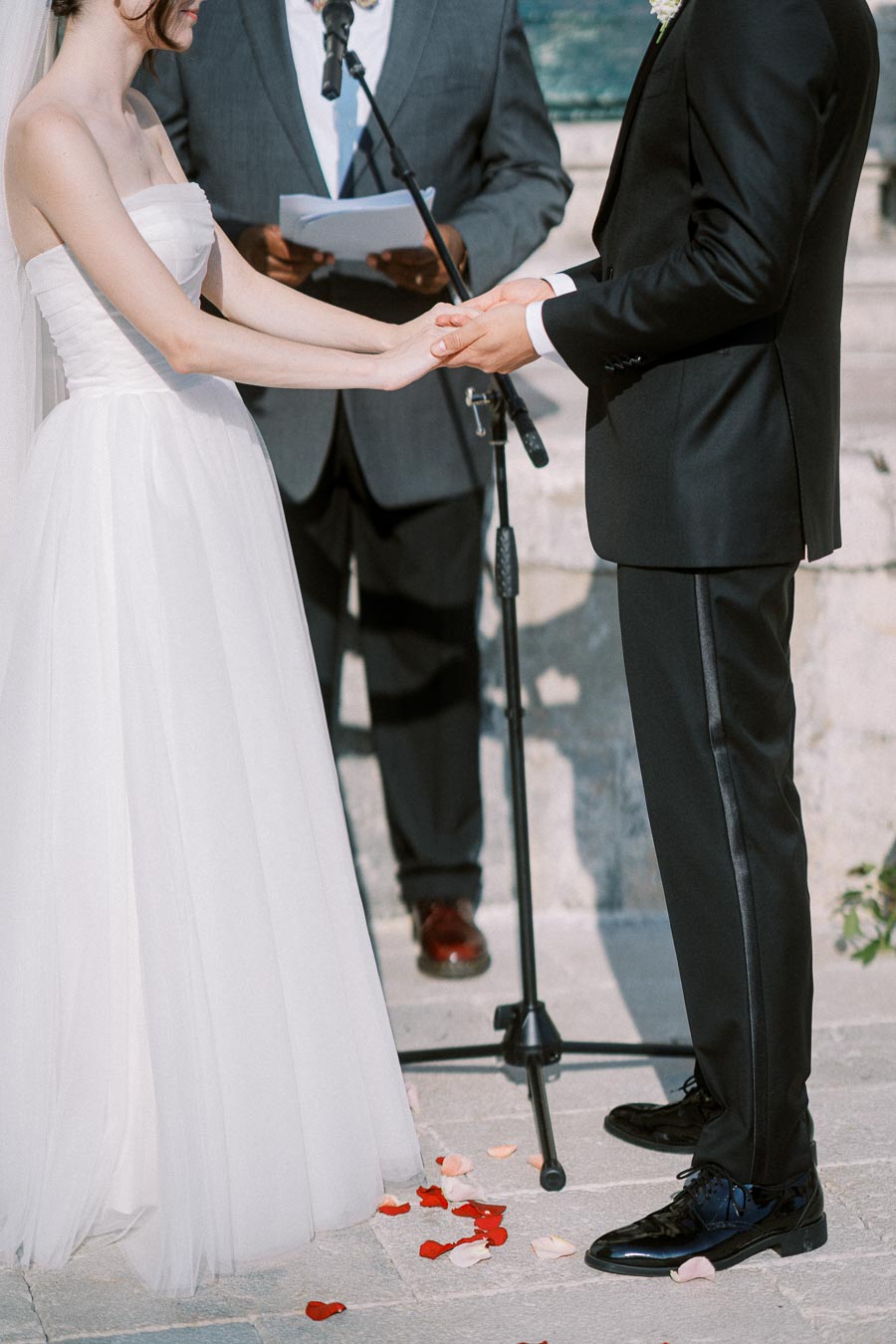A bride in a white wedding dress and a groom in a black suit hold hands during a wedding ceremony, with a person in formal attire officiating and rose petals scattered on the ground.