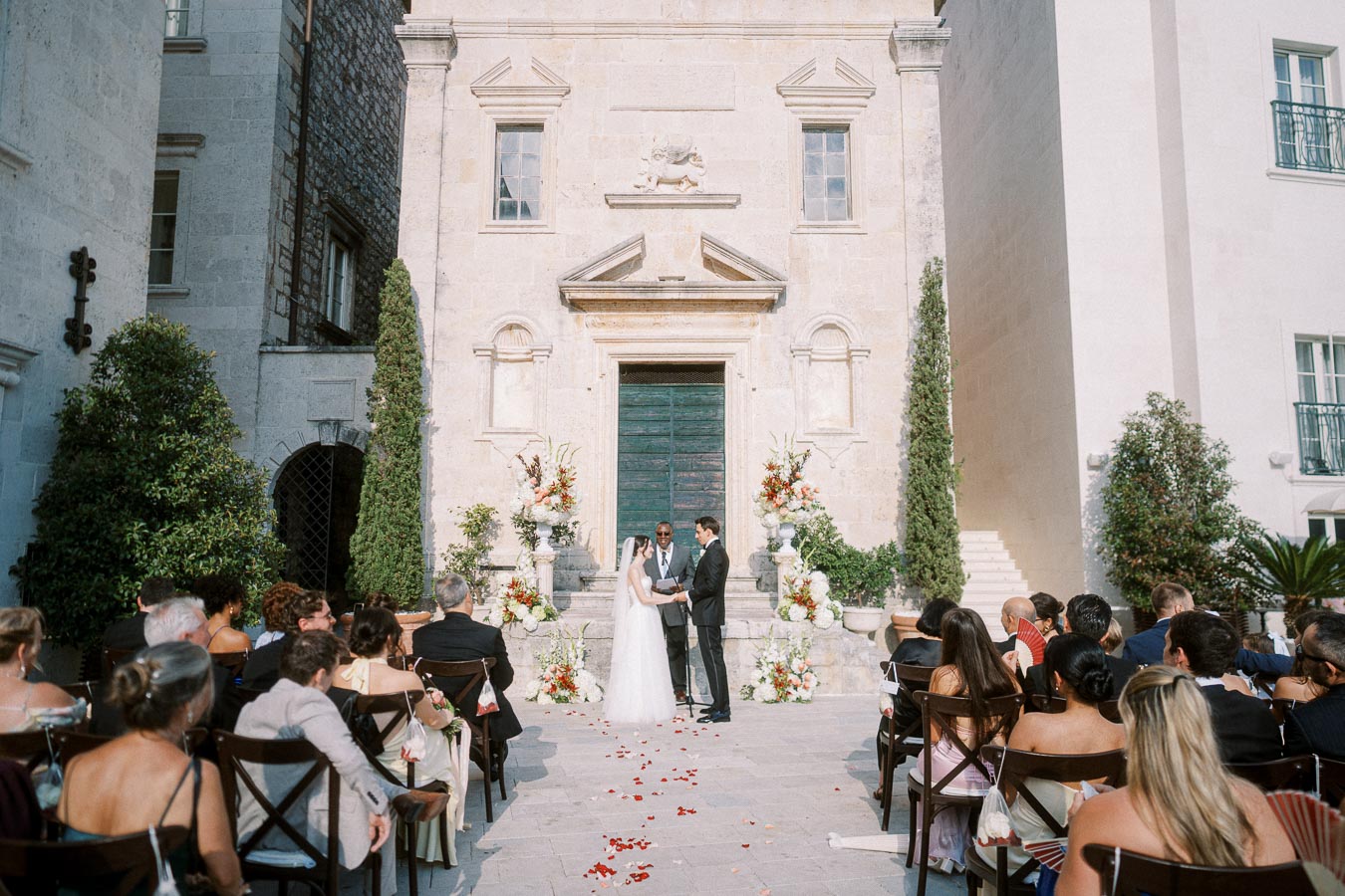 Outdoor wedding ceremony at historic stone venue with a couple exchanging vows in front of guests, surrounded by elegant floral arrangements and architecture.