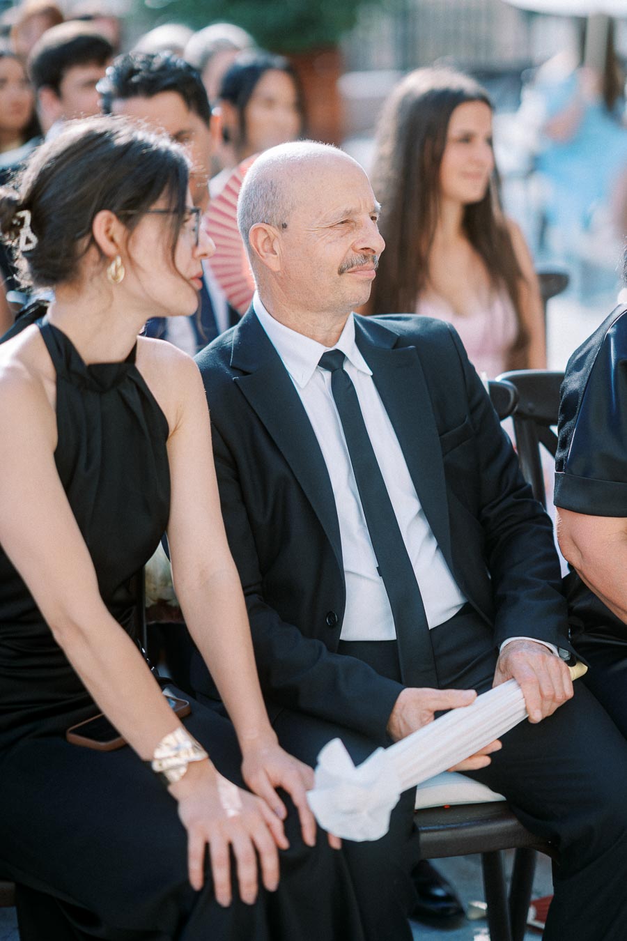 A man and woman in formal attire seated at an outdoor event, surrounded by other guests in a festive setting.