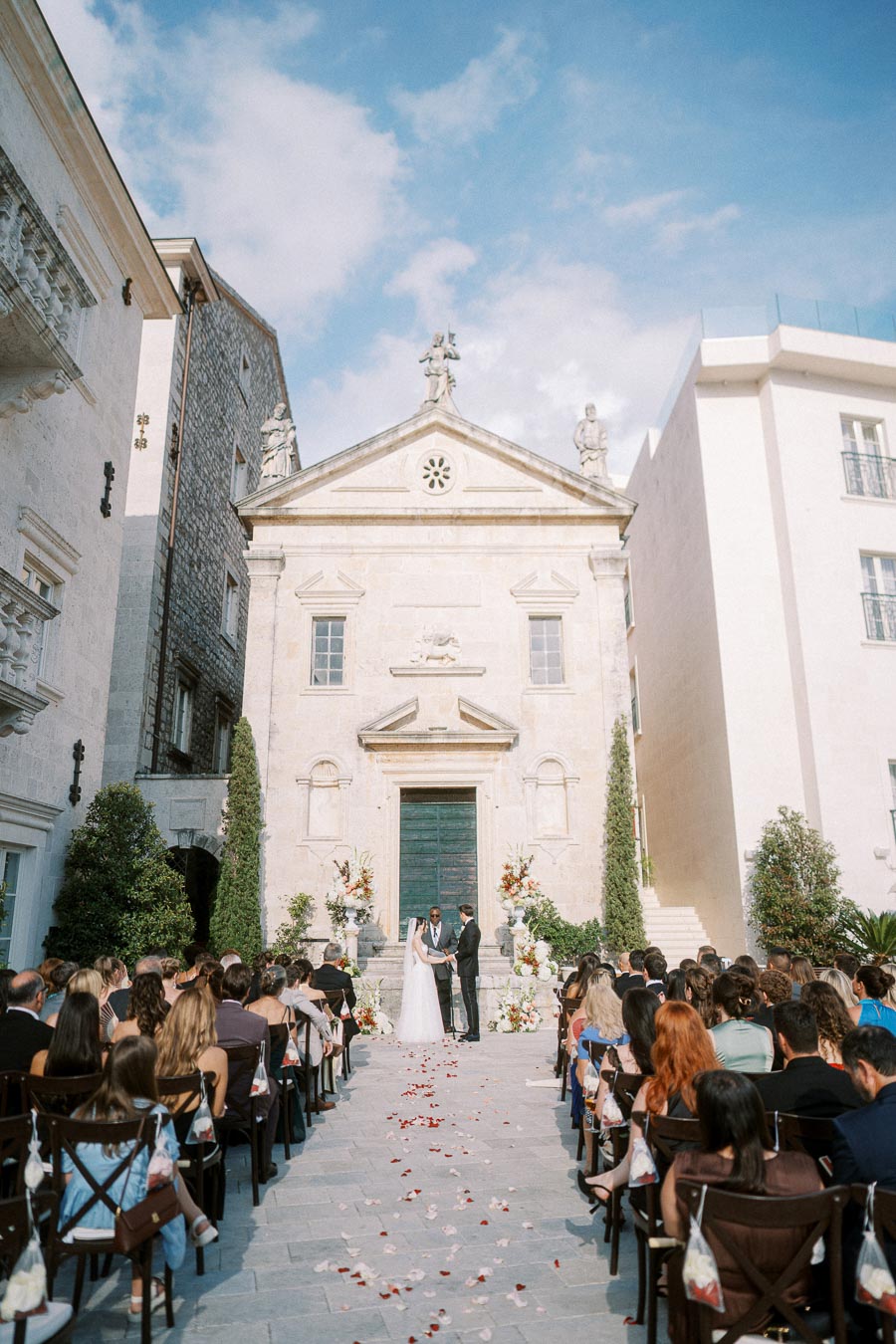 Outdoor wedding ceremony at a historic European church, featuring a bride and groom standing at the altar. The scene is set under a clear blue sky with guests seated on either side, surrounded by elegant floral arrangements.