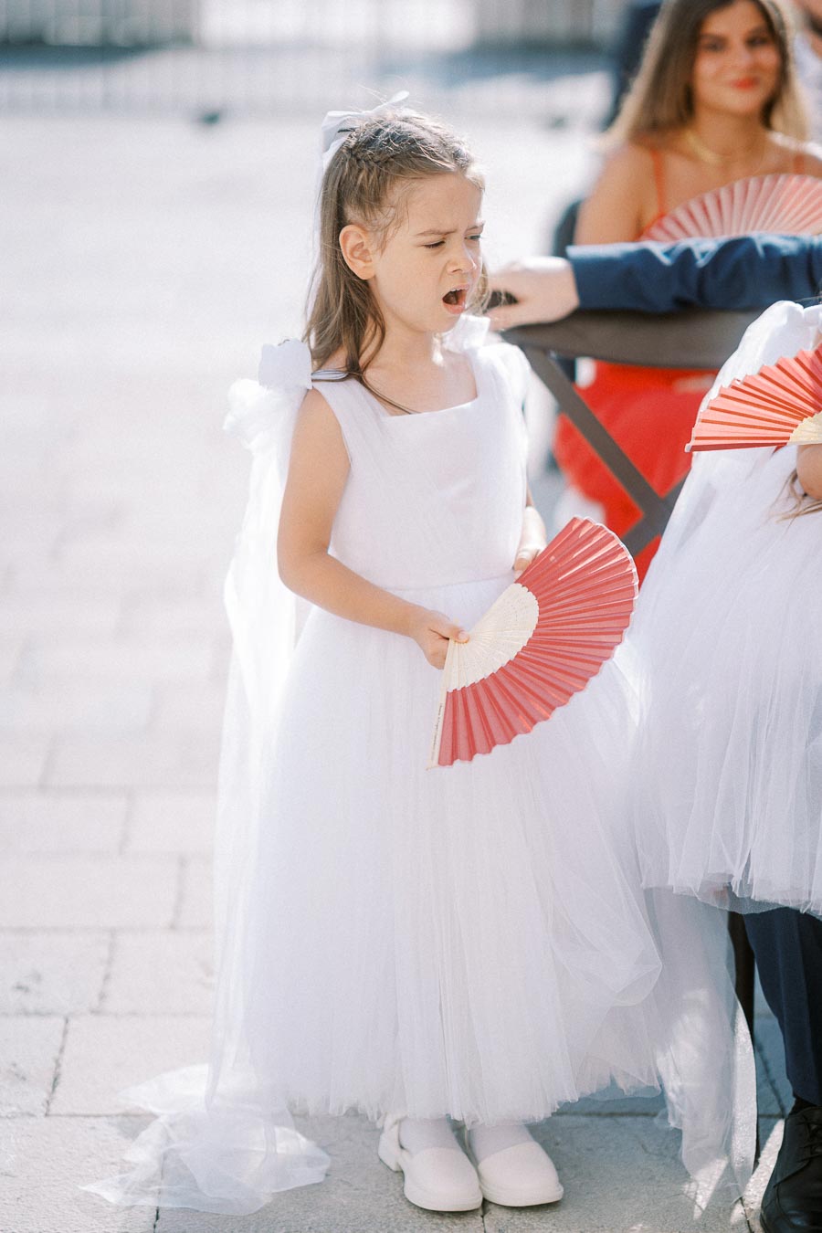 A young girl in a white dress holding a red and white fan, standing outdoors on a sunny day, with another person blurred in the background wearing a red outfit.